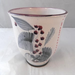 May include: A ceramic cup with a cream-coloured base featuring hand-painted grey foliage and clusters of small dark red berries. The rim is accented with a thin dark purple band, and a vertical line of small red dots runs down the side of the vessel.