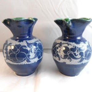 May include: Two blue ceramic vases with white floral patterns. The vases have a ruffled green rim and a bulbous body. The design features a repeating floral pattern around the middle of the vase.