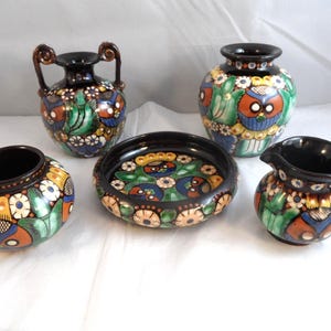May include: A set of five ceramic items featuring a floral and owl motif. The collection comprises a vase with handles, a small jug, a bowl, and two small vases. The pieces are decorated with orange, blue, green, and white floral patterns.