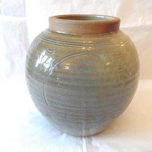 May include: A round ceramic vase with a glossy sage green glaze and a wide, unglazed terracotta-coloured rim. The body of the vase features subtle, incised swirling line patterns that wrap around the exterior, creating a textured surface.
