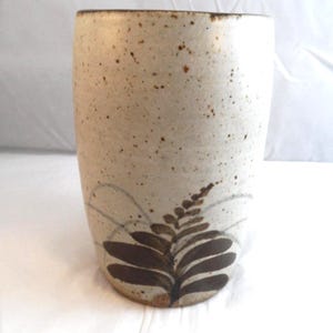 May include: A cylindrical ceramic vase in off-white with brown speckles. It has a hand-painted fern design in brown and grey. The rim is a darker brown. The vase is approximately 15 cm tall.