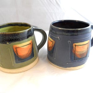 May include: Two ceramic mugs with rounded bodies and sturdy handles. The left mug is olive green with a dark rim, while the right mug is slate blue. Both feature a repeating pattern of orange and brown square motifs with thin white lines.