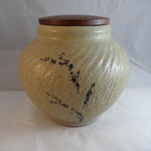 May include: A beige ceramic jar with a textured surface and a dark wooden lid. The jar features a decorative pattern of dark blue floral accents. The jar is a round shape.