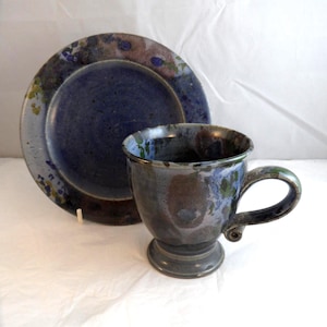 May include: A ceramic mug and matching plate featuring a mottled glaze in shades of deep blue, purple, and earthy brown. The mug has a pedestal base and a curved handle, while the plate displays a wide rim with speckled blue and green accents.