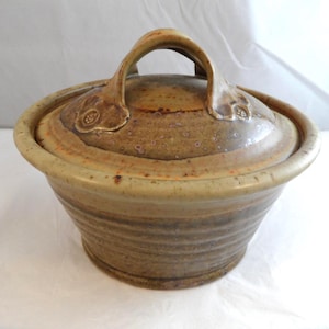 May include: A round, tan-coloured ceramic lidded bowl with a speckled glaze finish. The bowl features horizontal ridges along the exterior base and a curved handle on the lid, which is decorated with small, embossed floral patterns on each side.