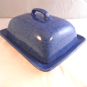 May include: A rectangular ceramic butter dish with a glossy finish in a deep blue colour featuring a speckled pattern throughout. The dish consists of a flat base with a raised edge and a matching domed lid with a curved handle on top.