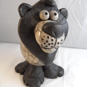 May include: A ceramic lion figurine featuring a matte black mane and paws, with a cream-coloured face and body. The body displays a dark crackle glaze pattern, while the face has two round eyes and a snout with small black dots for whiskers.