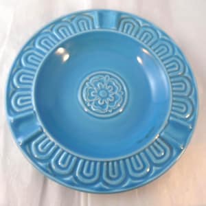May include: A round, bright blue ceramic ashtray featuring a central embossed floral medallion design. The outer rim is decorated with a repeating U-shaped pattern. The piece has a smooth, glossy finish and includes integrated notches along the edge.