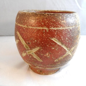 May include: A rounded ceramic bowl with a textured, reddish-brown exterior featuring carved, light-coloured cross-shaped markings. The vessel has a wide opening, a tapered base, and a speckled, matte finish across its entire outer surface.