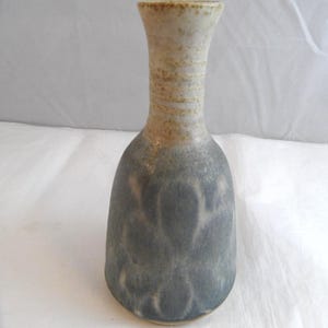 May include: A ceramic vase with a narrow neck and a wider, rounded base. The upper portion features a speckled beige finish with horizontal ridges, while the lower body displays a mottled, dark grey glaze with a subtle, cloud-like pattern.