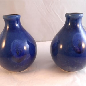 May include: A pair of matching ceramic vases with a rounded, bulbous body and a narrow neck. The vases feature a deep, glossy cobalt blue glaze with subtle, darker mottled patches and a smooth, reflective finish throughout the entire surface.