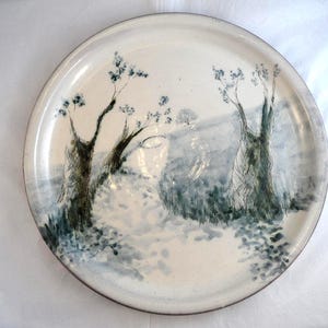 May include: A round ceramic plate with a cream-coloured base featuring a hand-painted landscape scene in shades of blue and black. The design depicts two prominent trees in the foreground with a path leading towards a distant hill and trees.