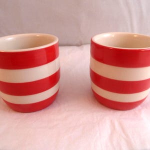 May include: Two ceramic cups featuring a horizontal striped pattern in bright red and off-white. Each cup has a rounded, handleless shape with a smooth, glossy finish and a wide, open top, displaying a consistent design across both items.
