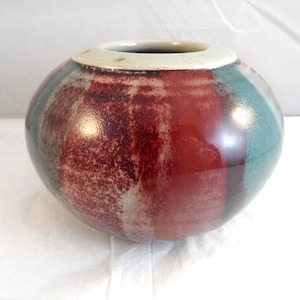 May include: A spherical ceramic vase with a wide opening and a cream-coloured rim. The vase is painted with vertical stripes of burgundy, teal, and light grey. The vase has a glossy finish and is likely used for decorative purposes.