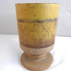 May include: A handcrafted ceramic goblet with a speckled yellow and brown glaze. The goblet has a wide, cylindrical top and a pedestal base. The color transitions from yellow to brown, with a speckled texture. The base is a lighter, unglazed colour.