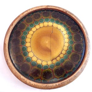 May include: A circular coaster with a wooden edge. The coaster has a central design in yellow and brown, encircled by a pattern of green, yellow, and brown shapes. The coaster is likely made of ceramic or a similar material, and is suitable for a cup or glass.