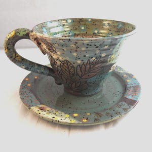 May include: A ceramic teacup and saucer set featuring a speckled blue-green glaze with dark brown leaf illustrations. The cup has a curved handle and a wide rim, while both pieces are decorated with small yellow dots and dark paint splatters.