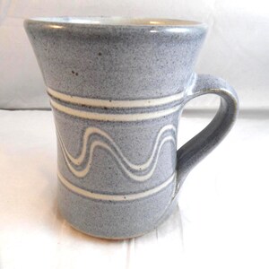 May include: A speckled light blue ceramic mug with a flared rim and a sturdy handle. The exterior features two horizontal white bands encircling the body, with a continuous white wavy line pattern positioned between the two bands.