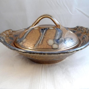 May include: A ceramic serving dish with a lid. The dish is a warm brown colour with blue and white accents. The lid has a handle and the rim of the dish has a decorative edge. The dish is likely handmade and has a rustic appearance.