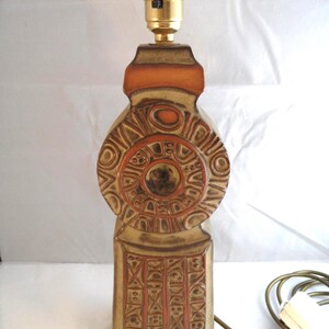 May include: A ceramic table lamp base featuring a tall, rectangular lower section with geometric relief patterns and a circular upper section with concentric ring designs. The lamp is finished in a matte tan glaze with burnt orange accents.