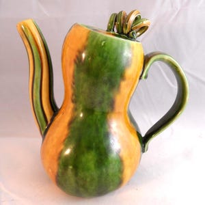 May include: A ceramic teapot with a distinctive gourd shape, striped with orange and green. The spout and handle are green, and the lid has a decorative swirl. The teapot is designed for serving tea.