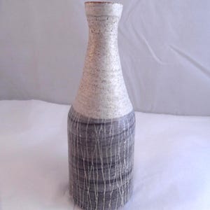 May include: A ceramic vase with a bottle-like form. The top is speckled white, leading to a grey base with vertical white lines. It has a narrow neck and a flared rim, displaying a minimalist design. The vase is approximately 25 cm tall.