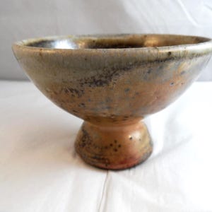 May include: A ceramic bowl with a pedestal base. The bowl has a wide, shallow shape with a speckled, earthy colour palette of brown, tan, and grey. The pedestal base is a similar colour and texture, with a rounded shape.