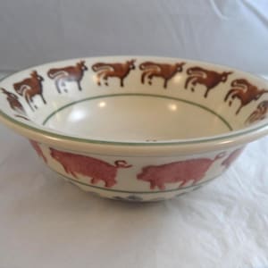 May include: A cream-coloured ceramic bowl featuring a hand-painted border of brown cows along the inner rim and a series of pink pigs encircling the exterior side. The bowl has a simple, rounded shape with a thin green line accenting the rim.