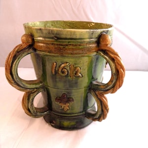 May include: A green ceramic vessel with two brown rope-like handles. The vessel has the number "1612" and a fleur-de-lis symbol embossed on the front. The rim and handles have a brown glaze.