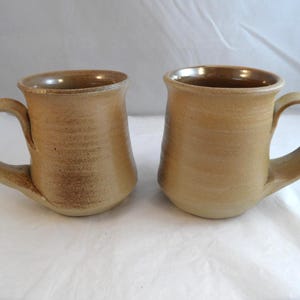 May include: Two matching ceramic mugs with a light tan, speckled finish. Each mug features a wide, rounded body, a thick curved handle, and a smooth, flared rim. The exterior surface displays subtle variations in colour and texture throughout.