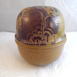 May include: A round, lidded ceramic container with a speckled tan base and a domed lid featuring a dark purple, abstract, organic pattern over a textured, mustard-yellow background. The vessel has a smooth, rounded silhouette and a visible seam.
