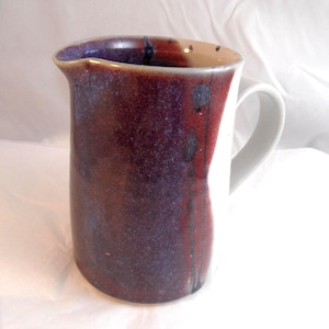 May include: A handcrafted ceramic jug with a white handle and a pouring spout. The jug features a deep purple and red glaze with dark speckles. The interior and handle are white, creating a striking contrast. Ideal for serving drinks.
