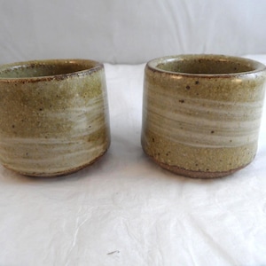May include: Two cylindrical ceramic cups featuring a speckled, earthy beige and olive green glaze with horizontal white bands. The cups have a thick, rounded rim and a textured, rustic finish with visible clay particles throughout the surface.