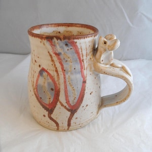 May include: A ceramic mug with a speckled cream glaze, featuring hand-painted abstract red and blue vertical shapes. The rim is accented with a brown band, and the handle is topped with a small, sculpted ceramic frog figure in a matching glaze.