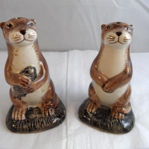 May include: A set of ceramic otter salt and pepper shakers. One otter holds a smaller otter. The otters are brown and cream-coloured with black eyes and sit on a dark grey base. The other otter has its paws crossed.