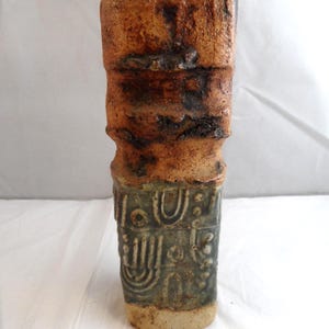 May include: A tall, rectangular ceramic vase with a textured surface. The upper portion features a mottled brown and dark rust-coloured glaze, while the lower section is a muted sage green with embossed geometric patterns and circular motifs.
