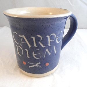 May include: A blue ceramic mug with a speckled finish and a white interior. The front features the text "CARPE DIEM" in white lettering, with two small orange dots and a white decorative symbol positioned below the text and above the base.