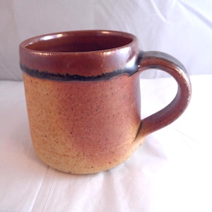 May include: A handcrafted ceramic mug with a speckled, rustic finish. The mug displays a colour gradient, shifting from a light tan base to a deep brown rim. A dark band accents the rim, and a curved handle completes the design.