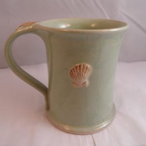 May include: A pale green ceramic mug with a handle and a seashell design. The mug has a cylindrical shape and a slightly flared rim. The seashell is a light tan colour and is attached to the mug's surface. The mug is approximately 10 cm tall.