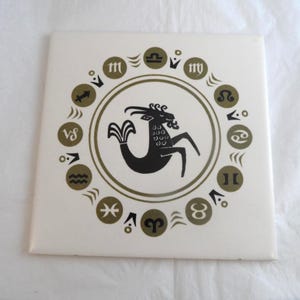 May include: A square white ceramic tile featuring a central black illustration of the Capricorn sea-goat zodiac symbol. Surrounding the centre is a circular border containing twelve olive green zodiac signs and small geometric accent marks.
