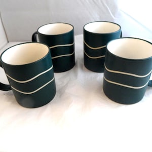 May include: A set of four dark green ceramic mugs, each featuring two horizontal, wavy white lines encircling the exterior. The mugs have a smooth, matte finish on the outside and a contrasting cream-coloured interior, with a sturdy handle on the side.