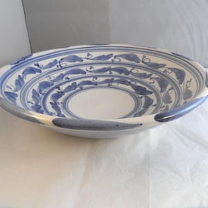 May include: A shallow, round ceramic bowl with a white base and hand-painted blue leaf and vine patterns arranged in concentric circles. The rim features a scalloped edge with alternating blue painted segments around the entire circumference.