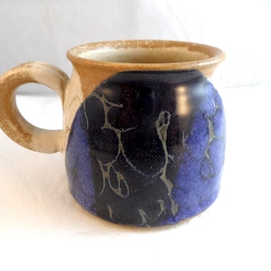 May include: A ceramic mug featuring a rounded body with a glossy dark blue and black glaze that has a crackled texture. The handle and the upper rim of the mug are finished in a contrasting matte beige, unglazed clay tone with a speckled appearance.