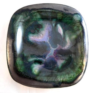 May include: A square ceramic dish with rounded corners featuring a glossy glaze in shades of deep black, forest green, and hints of violet. The centre displays a swirling, marbled pattern where the colours blend together into a smooth, reflective finish.