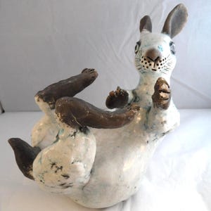 May include: A ceramic sculpture of a rabbit in a reclining position. The rabbit has a white body with blue crackle glaze accents and dark brown ears, paws, and feet. The sculpture features detailed facial markings and textured surfaces.