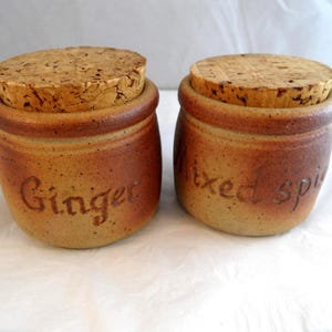 May include: Two round, speckled brown ceramic spice jars with cork stoppers. The left jar has the word Ginger embossed on the front, and the right jar has the words Mixed spice embossed on the front. Both jars feature a darker brown rim.