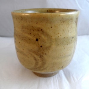 May include: A ceramic cup with a speckled tan and beige glaze. The exterior surface features subtle, wavy, horizontal indentations circling the body. The cup has a rounded, slightly tapered shape with a smooth rim and a distinct, unglazed base.