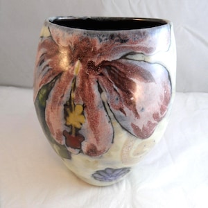 May include: A ceramic vase with a rounded, bulbous shape featuring a hand-painted floral design. The exterior displays a large, prominent pink and brown flower with a yellow centre, accented by smaller purple floral motifs against a cream background.