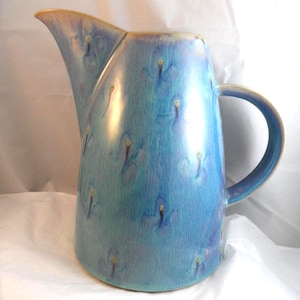 May include: A ceramic jug with a blue and turquoise glaze. The jug has a curved spout, a rounded body, and a handle. The glaze has a textured appearance with small, raised floral-like details. The rim and handle have a golden-yellow trim.