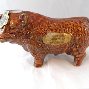 May include: A brown ceramic decanter shaped like a Highland cow with a textured coat. A gold label on the side reads RUTHERFORD'S SCOTCH WHISKY 5cl 40% vol. A small gold seal is attached to the top of the head between the ears of the animal figure.